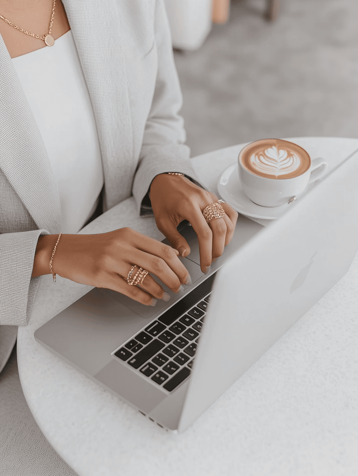 Person typing on a laptop with gold jewelry, next to a cappuccino on a white table. Modern and minimalistic workspace vibe.